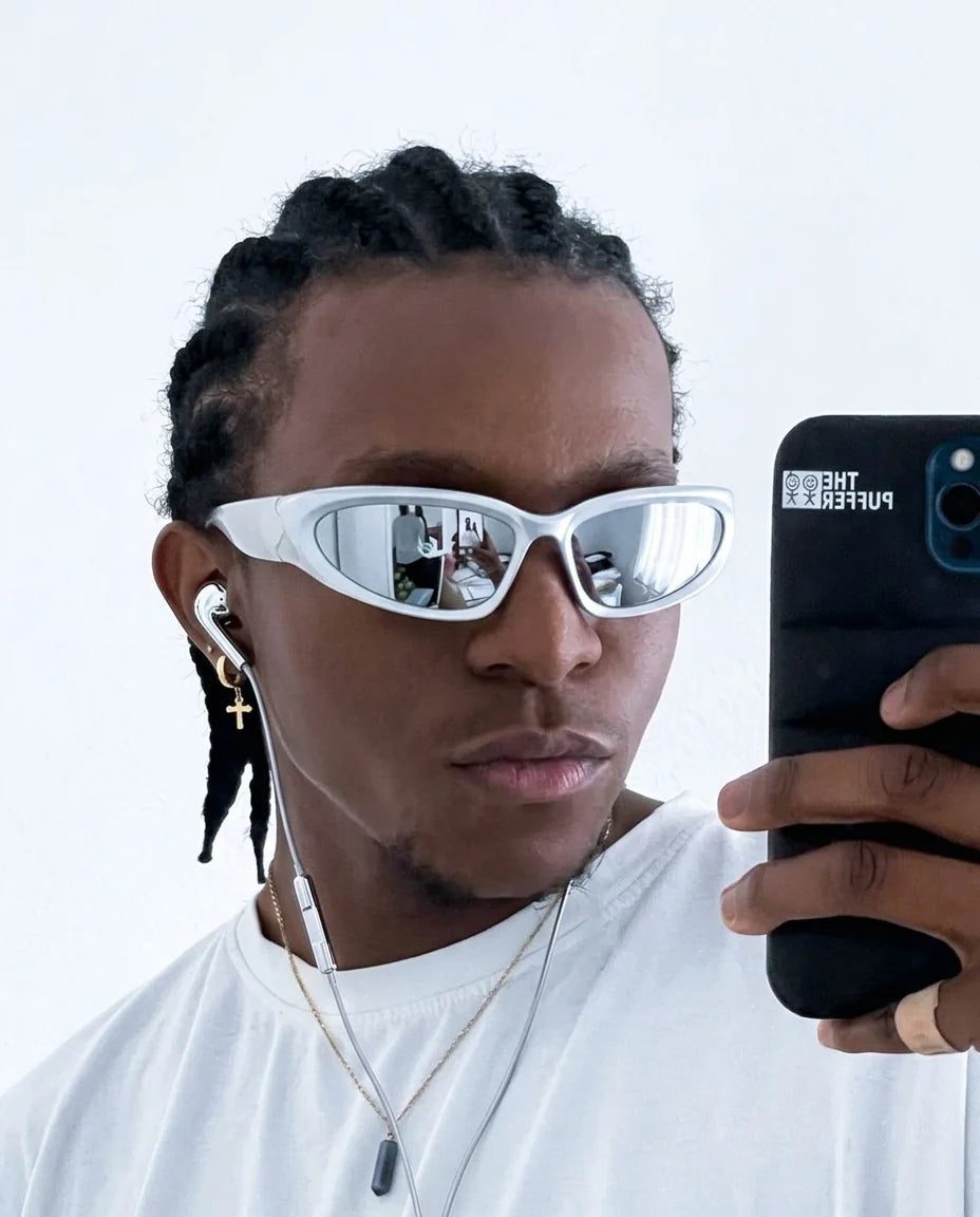 Man in chrome wraparound sunglasses taking a mirror selfie with a phone, wired earbuds in
