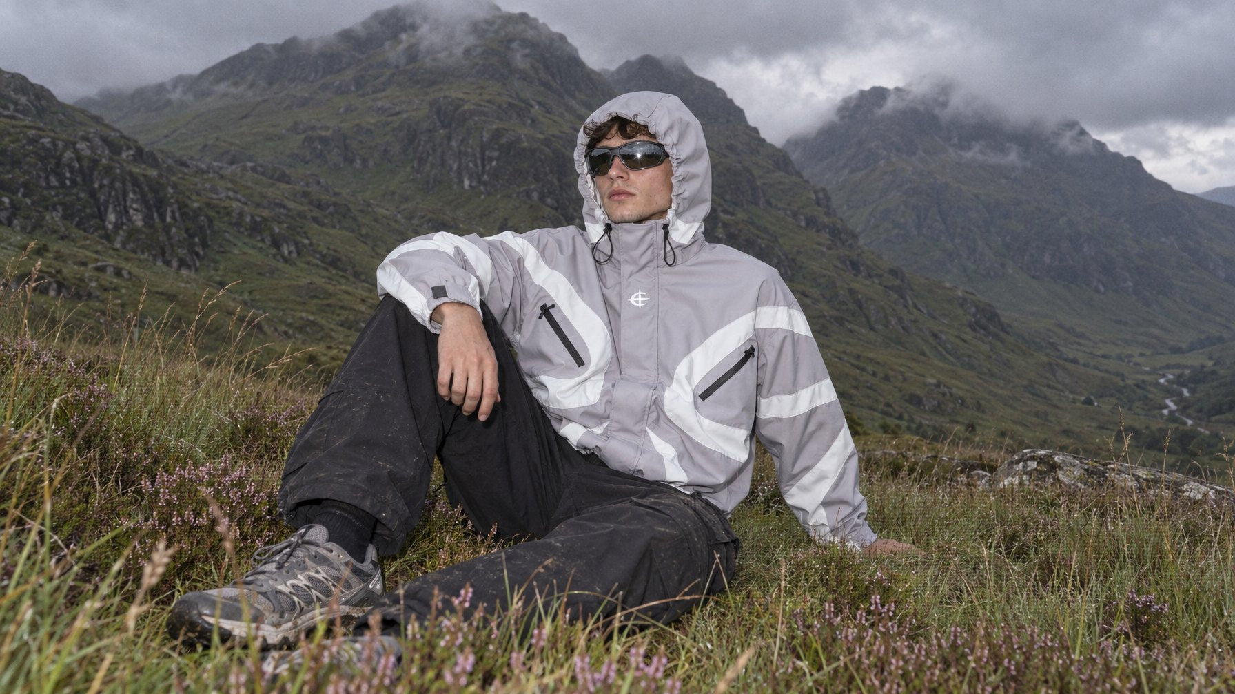 Man in the silver windbreaker with the hood up and wraparound sunglasses, sitting in heather in the Scottish Highlands with mountains and low cloud behind